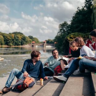 Eine Gruppe Studierender sitzt auf einer Treppe am Wasser, es ist sommerliches Wetter.