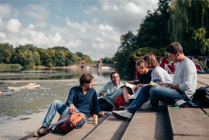 Eine Gruppe Studierender sitzt auf einer Treppe am Wasser, es ist sommerliches Wetter.