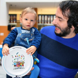 Ein Vater hat sein Kind auf dem Arm und lächelt es an. Das Kind hält einen leeren Kinderteller in der Hand.