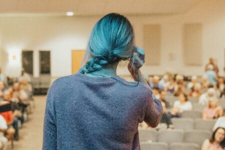 Eine Frau steht vor Menschen in einer Art Hörsaal. Sie fasst sich nervös ans Ohr. Man sieht sie von hinten.