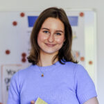 Portrait von Celina Bohmann. Sie steht vor einer Magnettafel und lächelt in die Kamera, in ihren Händen hält sie eine Mappe.