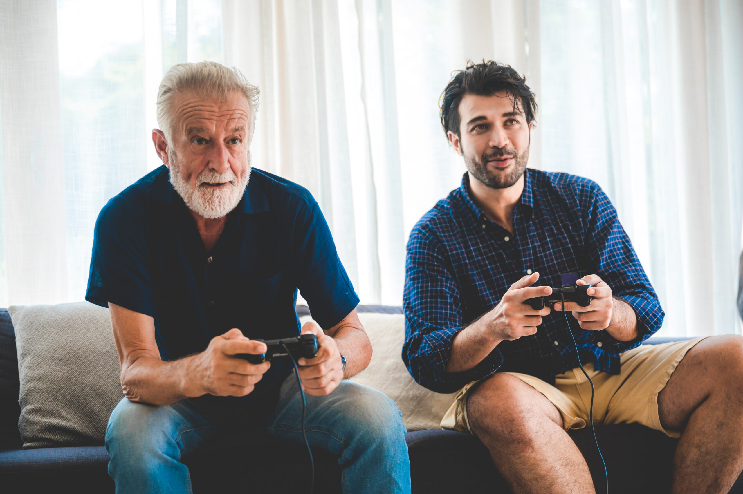 An older man and a younger man are sitting next to each other on the sofa, holding controllers in their hands and playing on a console.