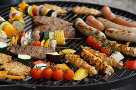 The photo shows the grill of a barbecue with sausages, grilled vegetables and meat on skewers, corn and tomatoes.