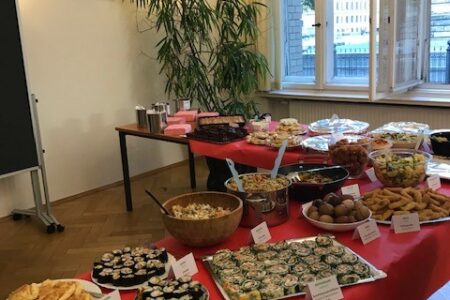 The picture shows a buffet with a variety of food such as sushi, fallafel or salads with description signs in a bright room.