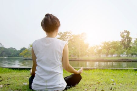 A person sits with his back to the viewer on a green meadow in front of a river and meditates in good weather.