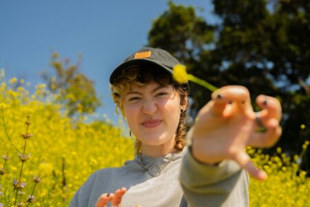 Das Bild zeigt eine junge Person mit Kappe und lockigen Haaren, die spielerisch eine gelbe Blume in die Kamera hält. Im Hintergrund blüht ein gelbes Blumenfeld unter blauem Himmel.