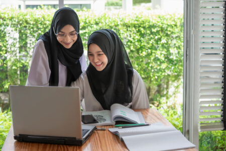 Zwei junge Frauen mit schwarzen Kopftüchern sitzen gemeinsam an einem Tisch vor einem Laptop und lächeln.