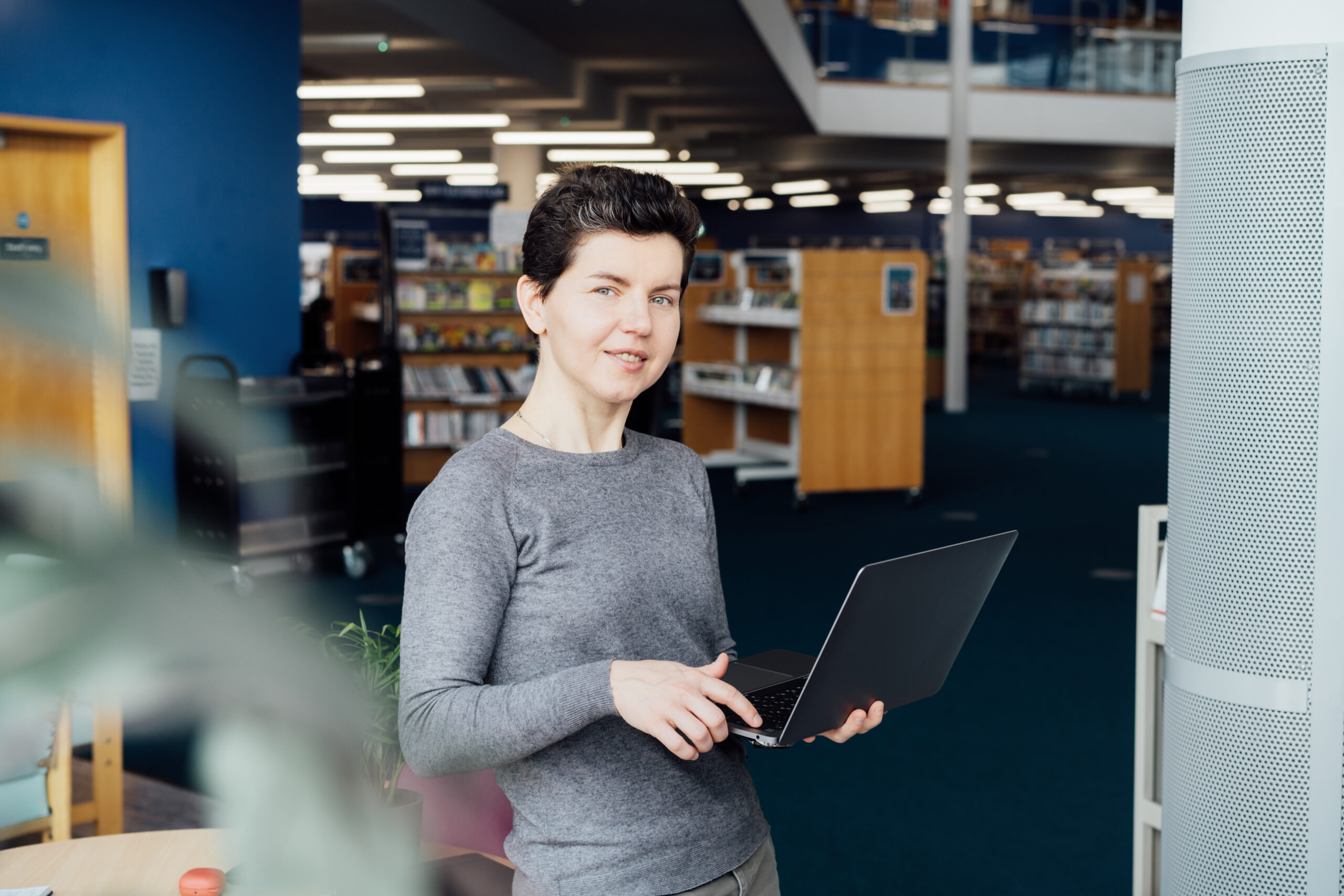Eine Person in einem grauen Pullover hält einen Laptop in einer Bibliothek.