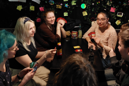 Gruppe an Studierenden sitzt in einer Kneipe um einen Tisch und spielt Karten, alle haben ein Getränk vor sich.