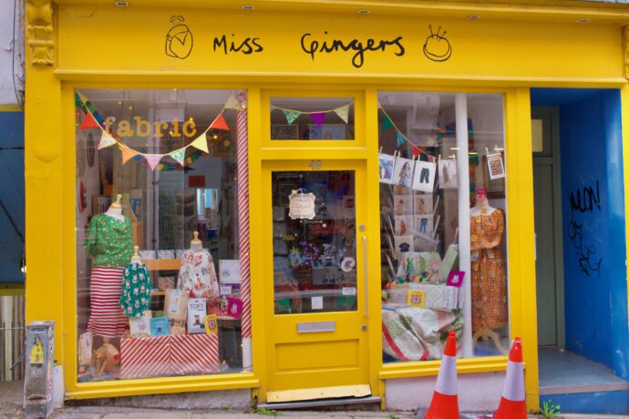 Bunte Fassade eines kleinen Ladens mit handgeschriebenem Schild „Miss Gingers“ und dekorierten Schaufenstern; zwei Verkehrshütchen davor.