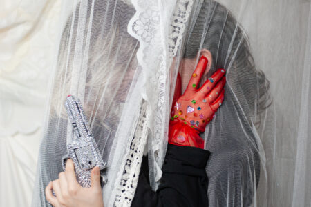 Zwei Frauen stehen hinter weißen Gardienen und küssen sich. Eine hat eine diamantbesetzte Pistole in der Hand, die andere trägt einen diamantbesetzten roten Handschuh.