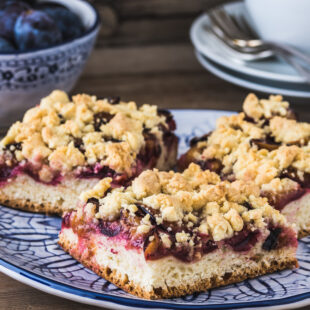 Auf einem blau-weißen Teller liegt Pflaumenkuchen, im Hintergrund stehen eine Schüssel mit Pflaumen und weiße Kaffeetassen.